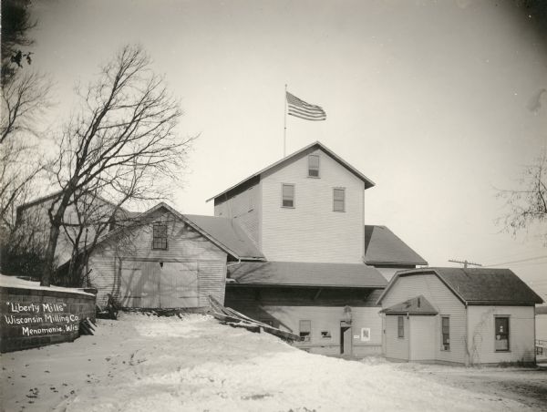 Caption reads: "'Liberty Mills' Wisconsin Milling Co. Menomonie, Wis." There is a flag on roof, and snow is on the ground.