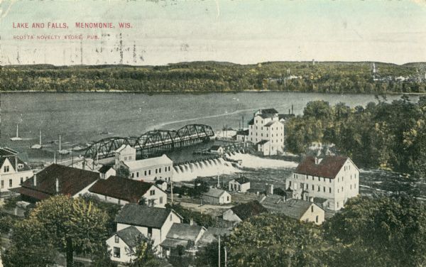 Elevated view across rooftops towards the dam on the Red Cedar River, also showing the electric power plant and mills. Caption reads: "Lake and Falls, Menomonie, Wis. Scotts Novelty Store, Pub."