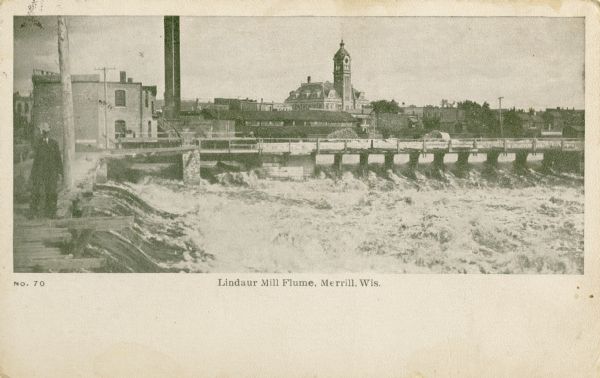 The Lindaur Mill Flume. A man is standing in the left foreground. In the distance is the Court House with clocktower. Caption reads: "Lindaur Mill Flume, Merrill, Wis."