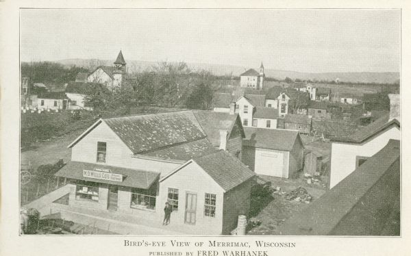 Elevated view of commercial buildings, houses, a church and a school in a neighborhood. In the foreground a person is standing at the side of a building with a sign that reads: "M.D. Wells Co's (?) Footwear". Hills are in the distance. Caption reads: "Bird's-eye View of Merrimac, Wisconsin. Published by Fred Warhanek."