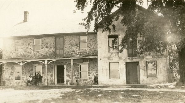 Exterior view of the Pheasant Branch Hotel or Stamm House. Men are sitting on the porch near the front door. A sign above the door reads, in part: "J. Yo....lood, Groceries."