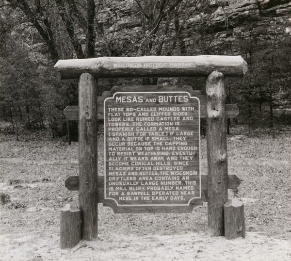 The Mesas and Buttes historical marker in Mill Bluff State Park. The marker reads: "Mesas and Buttes. These so-called mounds with flat tops and cliffed sides look like ruined castles and towers. The formation is properly called a mesa (Spanish for table) if large, and a butte if small. They occur because the capping material on top is hard enough to resist weathering. Eventually it wears away and they become conical hills. Since glaciers often destroyed mesas and buttes, the Wisconsin driftless area contains an unusually large number. This is Mill Bluff, probably named for a sawmill operated near here in the early days."