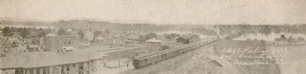 Elevated view of the town from a boxcar. A railroad train is headed towards a bridge over the lake. Caption reads: The ideal fishing resort of the One Thousand Lakes, Minocqua, Wis. 1916".