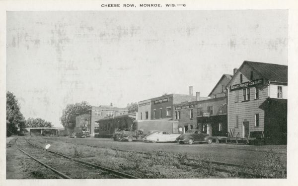 View across railroad tracks towards Cheese Row. Automobiles and trucks are parked near the buildings. Caption reads: "Cheese Row, Monroe, Wis."