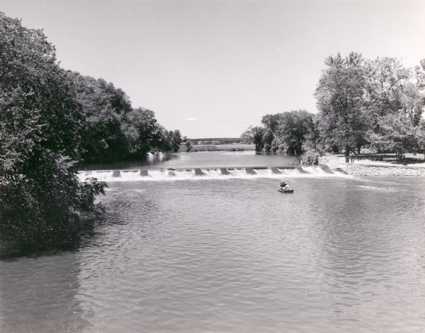 Slightly elevated view across water towards men fishing from a boat below the dam on the Fox River. Two other men are standing on the right shoreline near a stone wall at the dam.