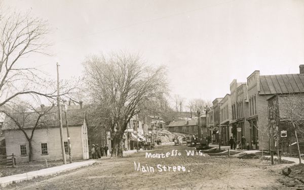 View down Main Street, with a hill at the end of the street. Pedestrians are walking on the sidewalks, and horse-drawn vehicles are along the unpaved street. Signs on the buildings along the right read: "Commercial House" and "City Market". A sign on a building on the left reads: "Millinery". Caption reads: "Montello, Wis., Main Street."