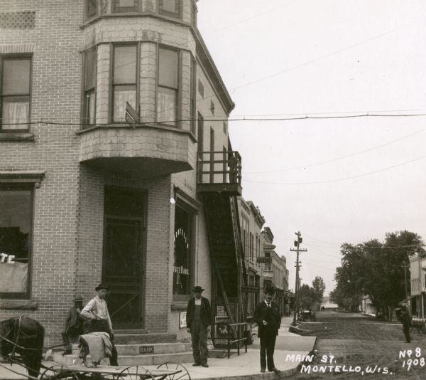 View from street towards men standing in front of bank at the corner of Main Street. A horse-drawn vehicle is in the foreground on the left. Signs along the sidewalk further down from the bank read: "Dr. T.F. Dempsey Dentist", "Metzler & Metzler Lawyers" and "[City] Market". Across the street on the right  is a sign for "Millinery". 