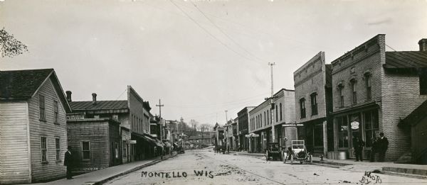 Panoramic view down Main Street, with a hill at the end. Pedestrians are on the sidewalks. Signs on the right side of the street read: "Garland Stoves and Ranges", "Commercial House" and "City Market". A sign on a storefront on the left reads: "Millinery".