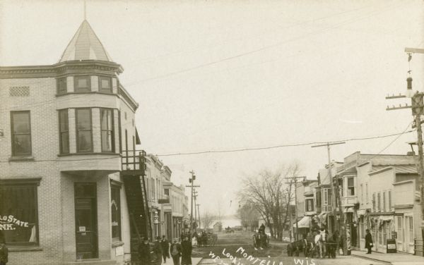 Storefronts and pedestrians on Main Street, with Buffalo Lake in the background. The Montello State Bank is on the left street corner. The City Market is further down the sidewalk. Caption reads: "Montello Looking West, Wis."
