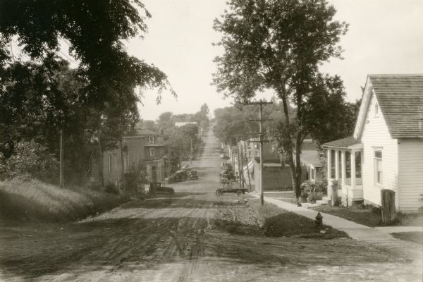 View down dirt road. Houses are on the right. Further down the road are commercial buildings and automobiles.
