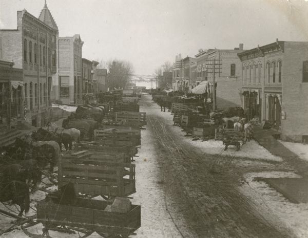 Elevated view of Main Street, looking north from the bridge. Horse-drawn wagons are parked on both sides of the snowy street.