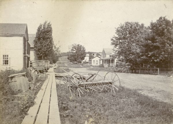 Main Street with plank sidewalk and dirt road. A piece of agricultural equipment is in the foreground.