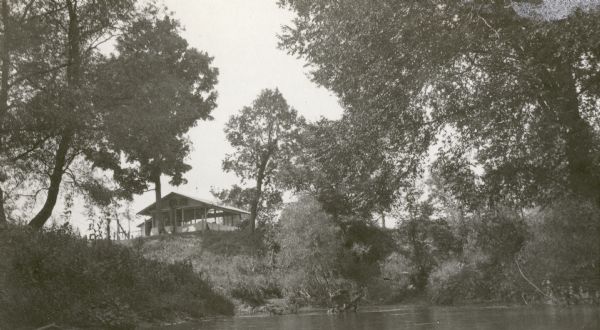View across water towards the C.R. Pickering Camp on Mill Creek, in summer. The shelter is on a hill overlooking the pond.