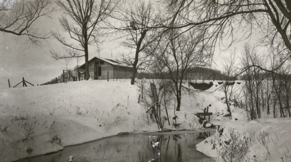 C.R. Pickering Camp on Mill Creek, in winter with snow on the ground. On the opposite shoreline is a shelter on the hill overlooking the creek, with a set of steps leading down to the water. Tree-covered hills are in the distance.