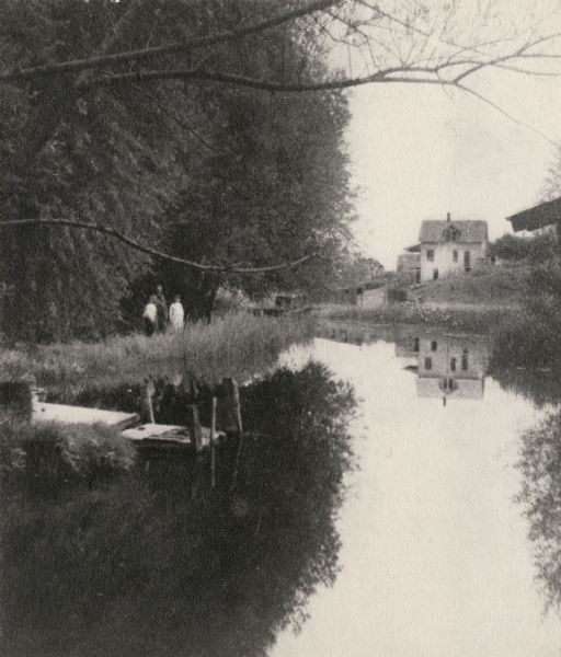 Nagowicka Roller Mills and mill race. Three people are standing on the opposite bank with the mill in the background.