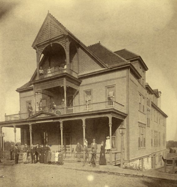 Necedah, Wisconsin. 1895-1899. Hotel Juneau with several people on front porch and balcony. On the middle balcony appear (the sequence is ambiguous) : Silas Reed, Mrs. Amelia Babcock Reed, Mrs. H.K. Reed, "Hattie," "Georgia" and Mrs. G.A. Potter. On the ground are C.E. Babcock (? left front) Mrs. J.W. Babcock (center), W.A. Reed (with dog) and (unidentified positions), Mr. and Mrs. J.M. Burch, "Eleanor," "Merrill" and "Homer."