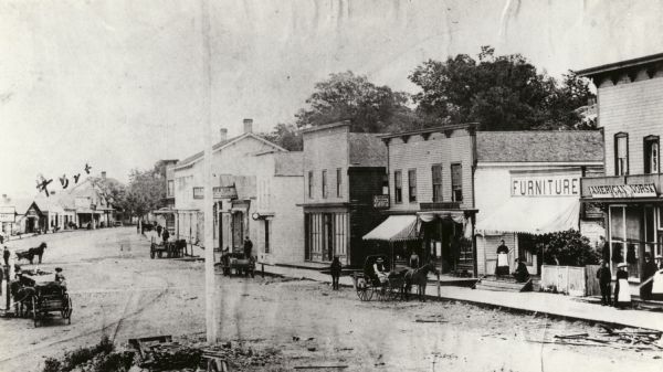 View of Main Street business district. Horses and buggies are on the street. Some business signs read: "American and Norsk Store," "Furniture," "L....re & Son, Dry Goods and Merchandise."