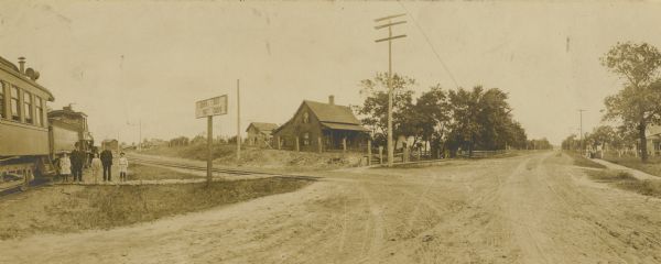 View of crossroads where the street meets the railroad tracks. Adults and children are standing to the left next to a train.