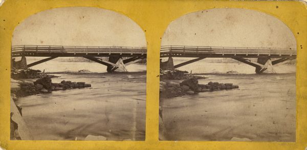 Stereograph view of Cedar Street Bridge(?) with a rocky shoreline on the left. The construction of this bridge is the same as the Cedar Street bridge, but as other landmarks are not visible, it is not definitely verified. Published by C.B. Manvill, Neenah, Wis. (variously spelled Manville).