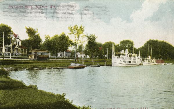 View from shoreline towards a large boat, perhaps an excursion boat, and sailboats docked further down along the curving shoreline. Beyond the boats is a trolley car running along the street, and houses are in the background. Caption reads: "Riverside Park, Neenah, Wisconsin."