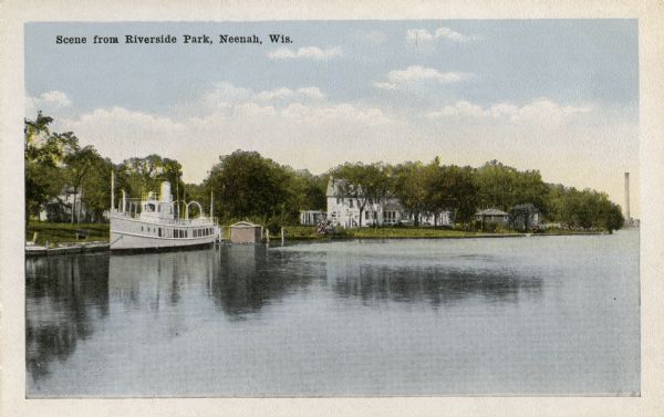 View across water towards an excursion boat moored at the shoreline of Riverside Park. There are houses beyond, and in the distance is a smokestack. Caption reads: "Scene from Riverside Park, Neenah, Wis."