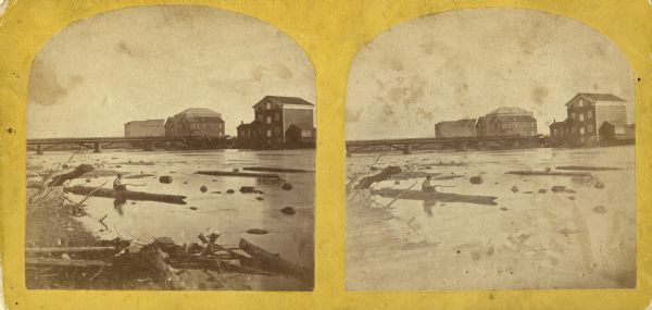 Stereograph view of the flour mill district. The Cedar Street Bridge is in the background, and the mills are, right to left going back, the Winnebago Flour Mills, Patten's Flour Mills, Island Cty Mills, Keystone Mills and the Atlantic Mills (Neenah Stone Mils in 1870). Since the mill to the extreme left is definitely labeled as the Atlantic Mills in the photograph, it would prove the date is not exactly 1870, but it cannot be much later as the buildings, bridge, etc. are exactly the same as pictured in an 1870 view. There is a young boy sitting on log in foreground (fishing?).