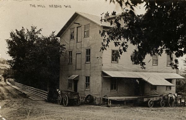 Two horse-drawn carts are parked near the mill. One horse-drawn cart is parked near a loading dock under a roof. On the left the road goes over a bridge, with a sidewalk and fence along the right side. Caption reads: "The Mill, Neosho, Wis."