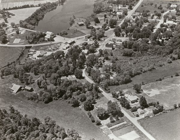 Aerial view looking north-northwest. View includes houses, roads, farm buildings, tractor-trailors (parked), other buildings.