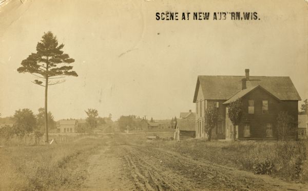 View down dirt road with a tree on the left, and a building (house?) on the right. More buildings are further down the road. Caption reads: "Scene at New Auburn, Wis."