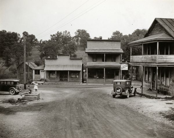Slightly elevated view of storefronts on Main Street. Sign on building reads: "Pabst Blue Ribbon Beer." Child standing at pump (water pump?) Two cars parked on street.