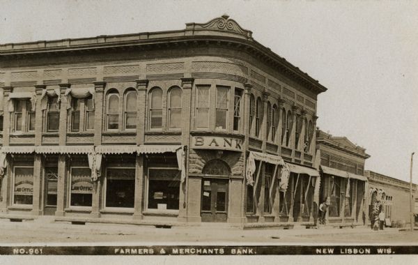 View of front of Farmers & Merchants Bank on a street corner. Other businesses in view include a law office and bakery. Three men are standing in front of the building. Caption reads: "Farmers & Merchants Bank. New Lisbon, Wis."