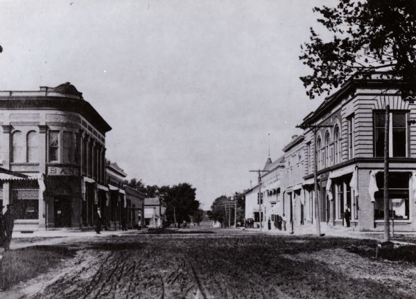 View down unpaved street. There is a bank on the corner on the left, and people are standing on the sidewalks.