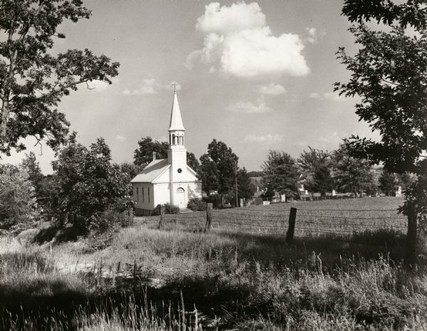 Exterior view across fence and field towards the Lutheran church and cemetery of a German or Norwegian settlement near the intersection of Highway 16 and County Highway O.
