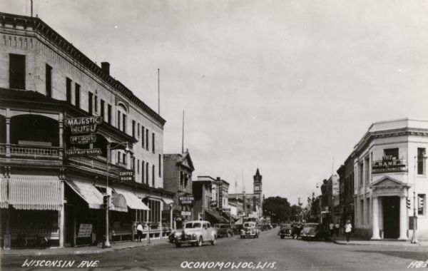 Street with retail shops, and automobiles along the avenue. Caption reads: "Wisconsin Ave., Oconomowoc, Wis." Some of the signs on the commercial buildings read: "Majestic Hotel, ...tap room, Pabst Blue Ribbon"; "Coffee room"; "Western Union"; "The Bank of Oconomowoc."