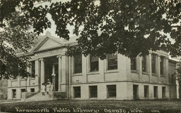 Exterior view of the Farnsworth Public Library. Four people are standing on front stepss (woman and girls).
