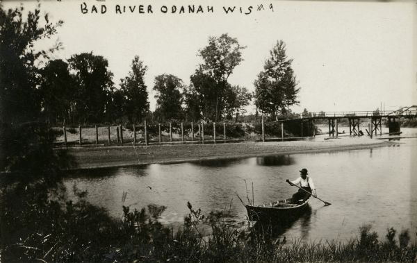 Man in canoe close to shoreline with bridge in background.