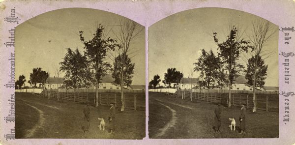 Stereograph. Probably the Presbyterian Mission School grounds on the Chippewa Indian reservation. Two boys and a dog are standing in the foreground, with a school building in the background.