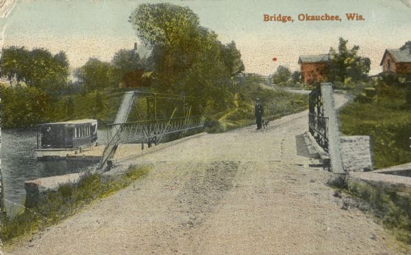 Man and dog standing near bridge. Buildings in background. There is a boat on the left on the water near the bridge. Caption reads: "Bridge, Okauchee, Wis."