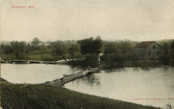 Elevated view of lake and and a bridge road. A man is standing on the road, and a boat is pulled up near the bridge. There is a building on the opposite shoreline. Caption reads: "Okauchee, Wis."