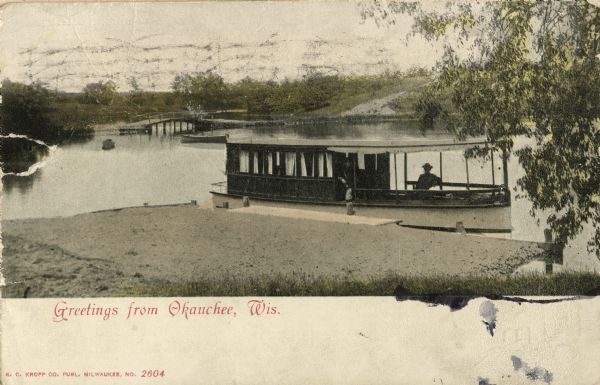 View from shoreline towards a man on a boat on lake, and a bridge road in the background on the left. Caption reads: "Greetings from Okauchee, Wis."