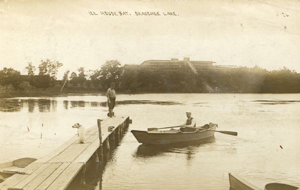 Ice House Bay and the lake. A man and a dog are standing on dock in the foreground, and on the right is a man in rowboat. There is a large  building on the opposite shoreline which has a long platform that is attached to the roof and leads down to the lake. Caption reads: "Ice House Bay, Okauchee Lake."