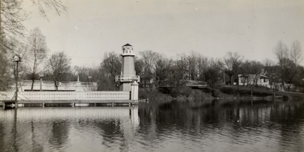 Lake Okauchee lighthouse. Buildings are in the background along a tree-covered shoreline.