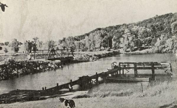 Bridge boat landing, with a dog standing in the foreground near a shoreline with a pier and a boat. There is a bridge across the water on the left, and tree-covered hills are in the background.