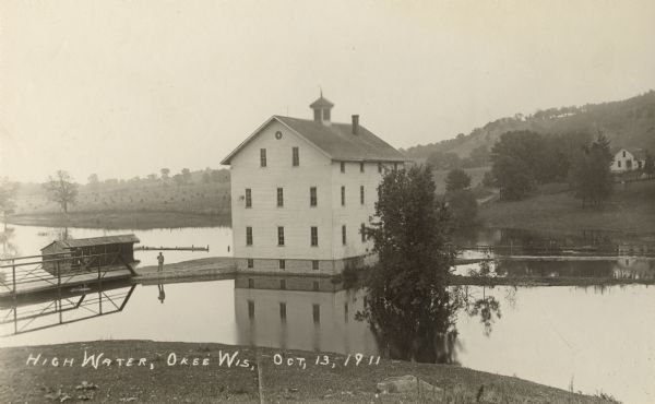 Elevated view of a mill during a flood, with a man standing on the left between a bridge and the mill. A house is on the side of a tree-covered hill on the right.