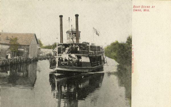 View towards the front of the excursion boat "Thistle" loaded with riders on the Fox River. Two people are standing on the right above the pilings near a building. Trees are on the right. Caption reads: "Boat Scene at Omro, Wis."