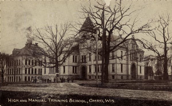 View across road towards a group of children standing in front of school buildings. Caption reads: "High and Manual Training School, Omro, Wis."