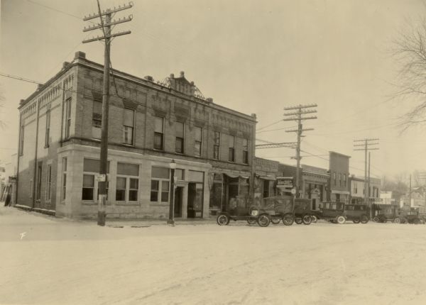 View across Main Street towards buildings on the left. In the center is Shurtleffs Ice Cream, Cigars, and Candy Shop. Automobiles are parked at an angle along the curb.