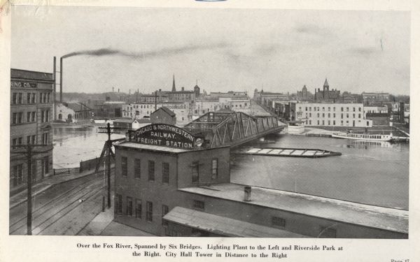 Elevated view of the Fox River Bridge. The building in the foreground has a sign that reads: "Chicago & Northwestern Railway. Freight Station." Caption reads: "Over the Fox River, Spanned by Six Bridges. Lighting Plant to the Left and Riverside Park at the Right. City Hall Tower in Distance to the Right".