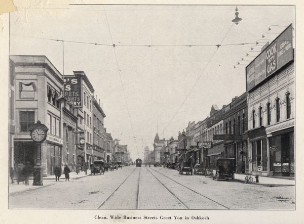 View down center of a street. Caption reads: "Clean, Wide, Business Streets Greet You in Oshkosh". On the left side of the street is a large clock on the sidewalk, and a sign for the "Hay Hardware Co." On the right is a billboard with advertisements for flour and coal.