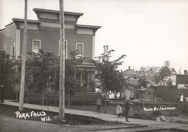 View across road towards the Park Hotel, with children playing on the sidewalk in front of the hotel. Caption reads: "Park Falls, Wis."